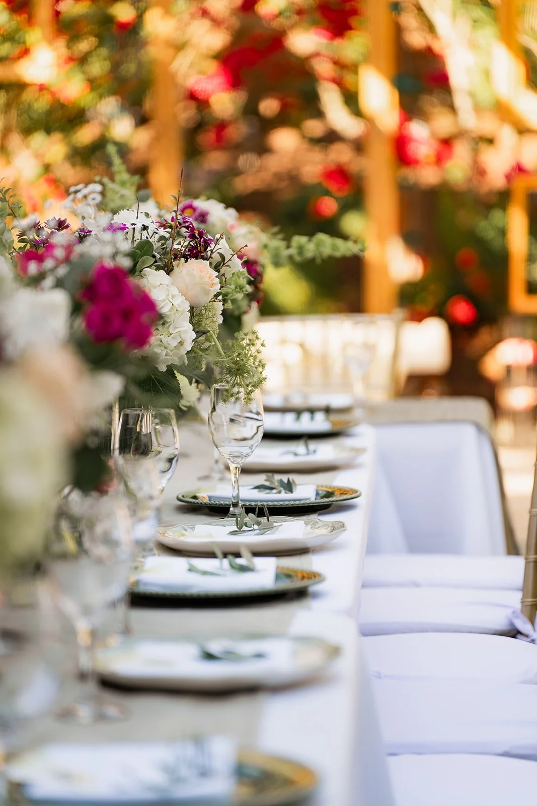 Elegant outdoor table set with white tablecloth, floral centerpieces, plates, silverware, and glasses, set for a formal event or wedding celebration.