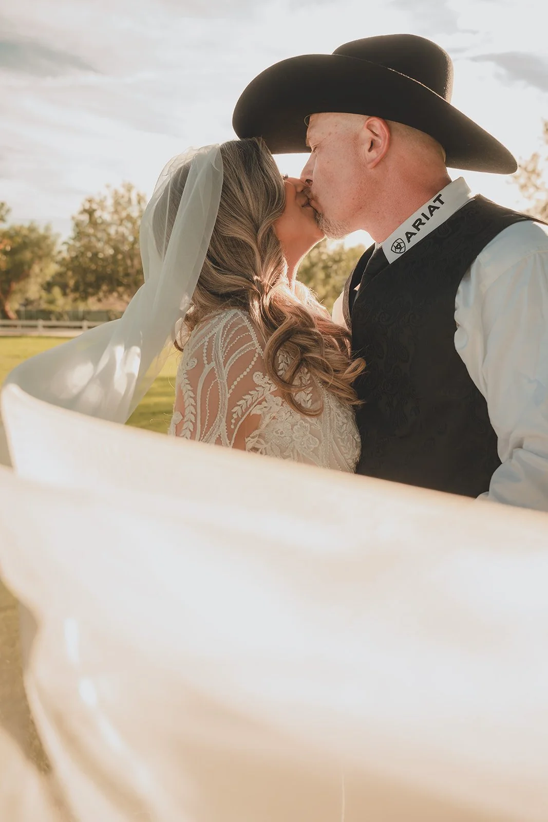 A bride and groom sharing a kiss outdoors during sunset, with the bride wearing a veil and lace wedding dress, and the groom in Western attire with a cowboy hat.