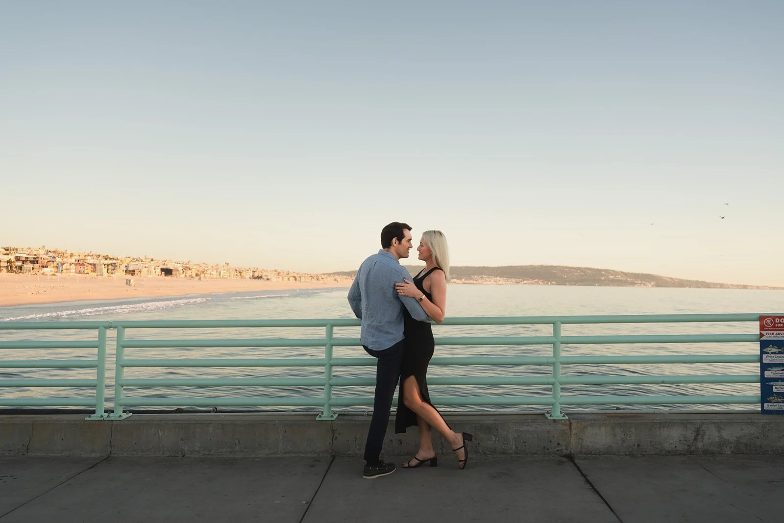 A couple standing close and facing each other on a pier, with the ocean and a distant shoreline in the background during sunset.