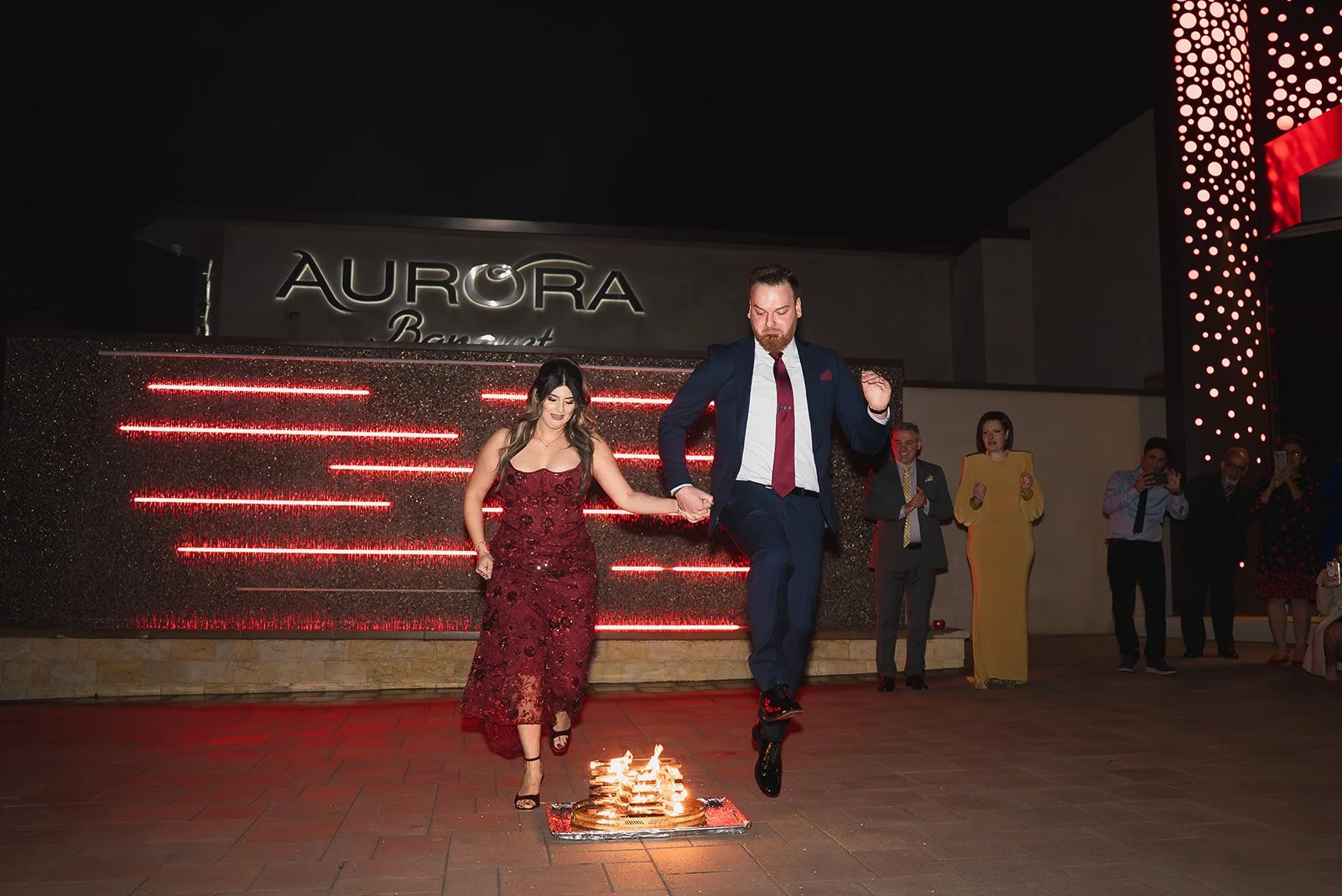 A couple dancing at an event with a small fire on the floor, surrounded by guests cheering. The venue is outdoor at night and has a sign that says "Aurora".