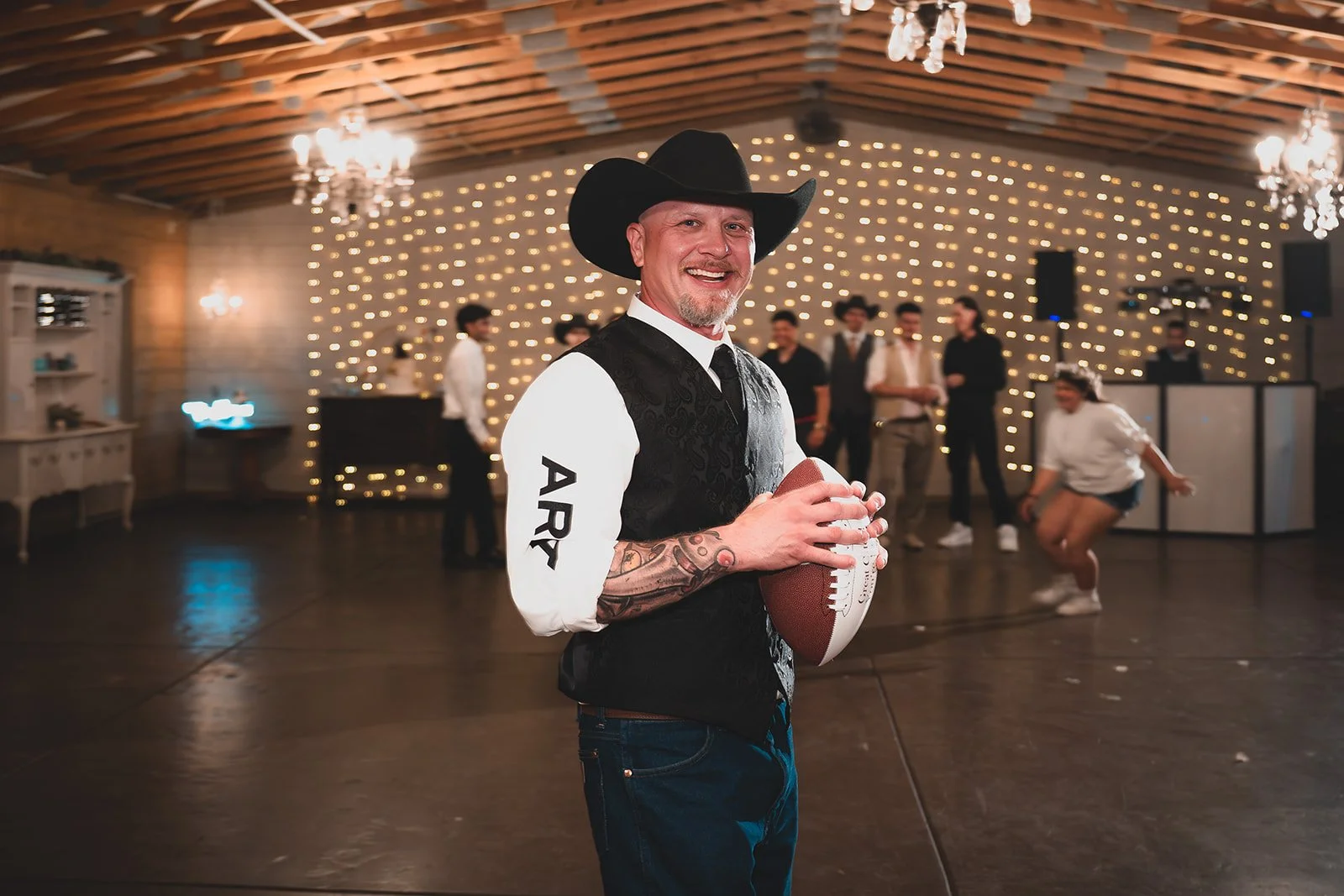 A man wearing a cowboy hat and a vest holding a football, smiling, at a party with a decorated backdrop and group of people in the background.