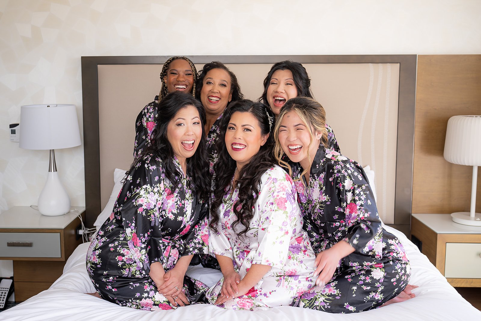 A group of seven women in floral robes sitting and kneeling on a bed, smiling and laughing together in a hotel room.