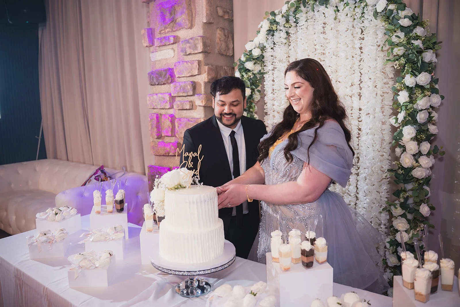 A bride and groom cutting a wedding cake at their celebration, surrounded by desserts and floral decorations.