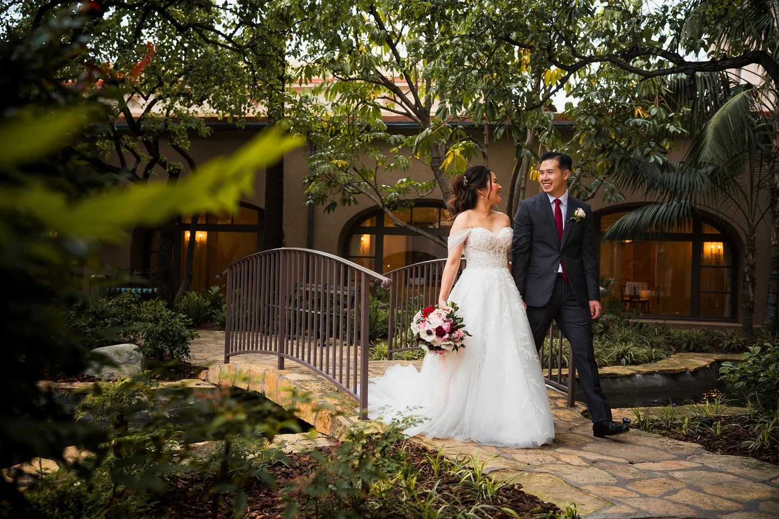 A bride and groom walking together on a stone pathway in a garden, smiling and looking at each other, with trees and a building with large windows in the background.