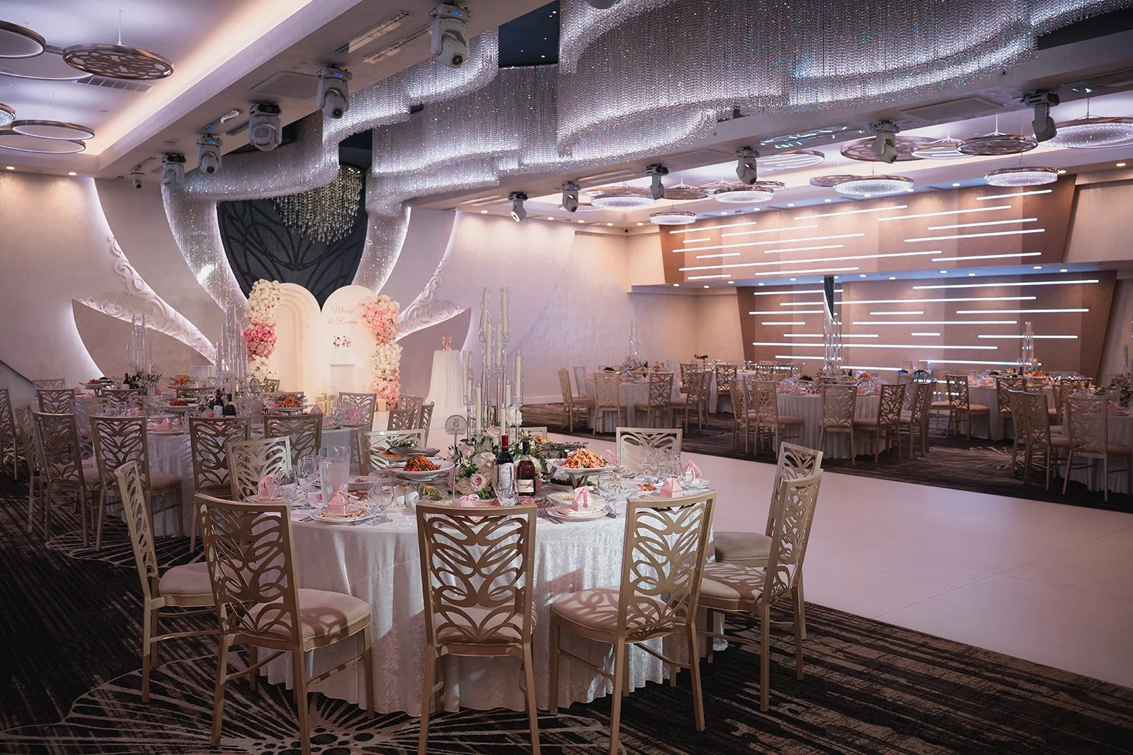 Elegant wedding reception hall with decorated round tables, floral centerpieces, and a backdrop with flower arrangements and a wedding arch.