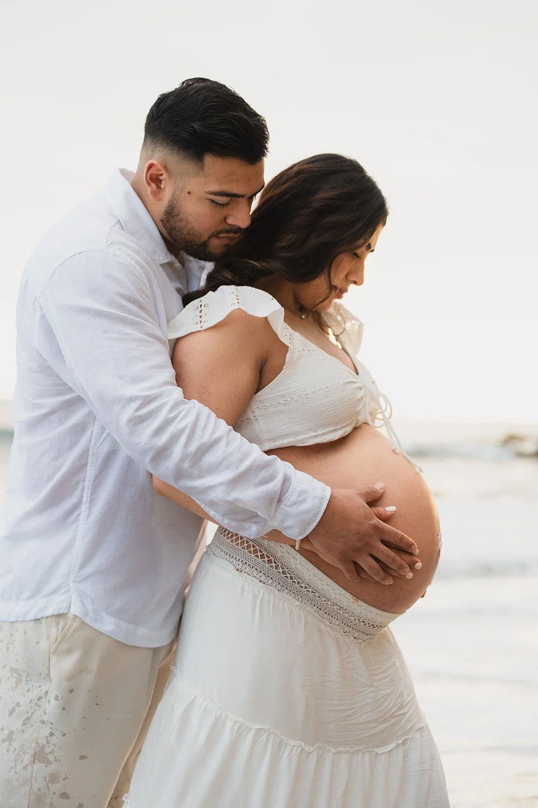 A man and pregnant woman on the beach, with the man holding her belly and both looking down gently.