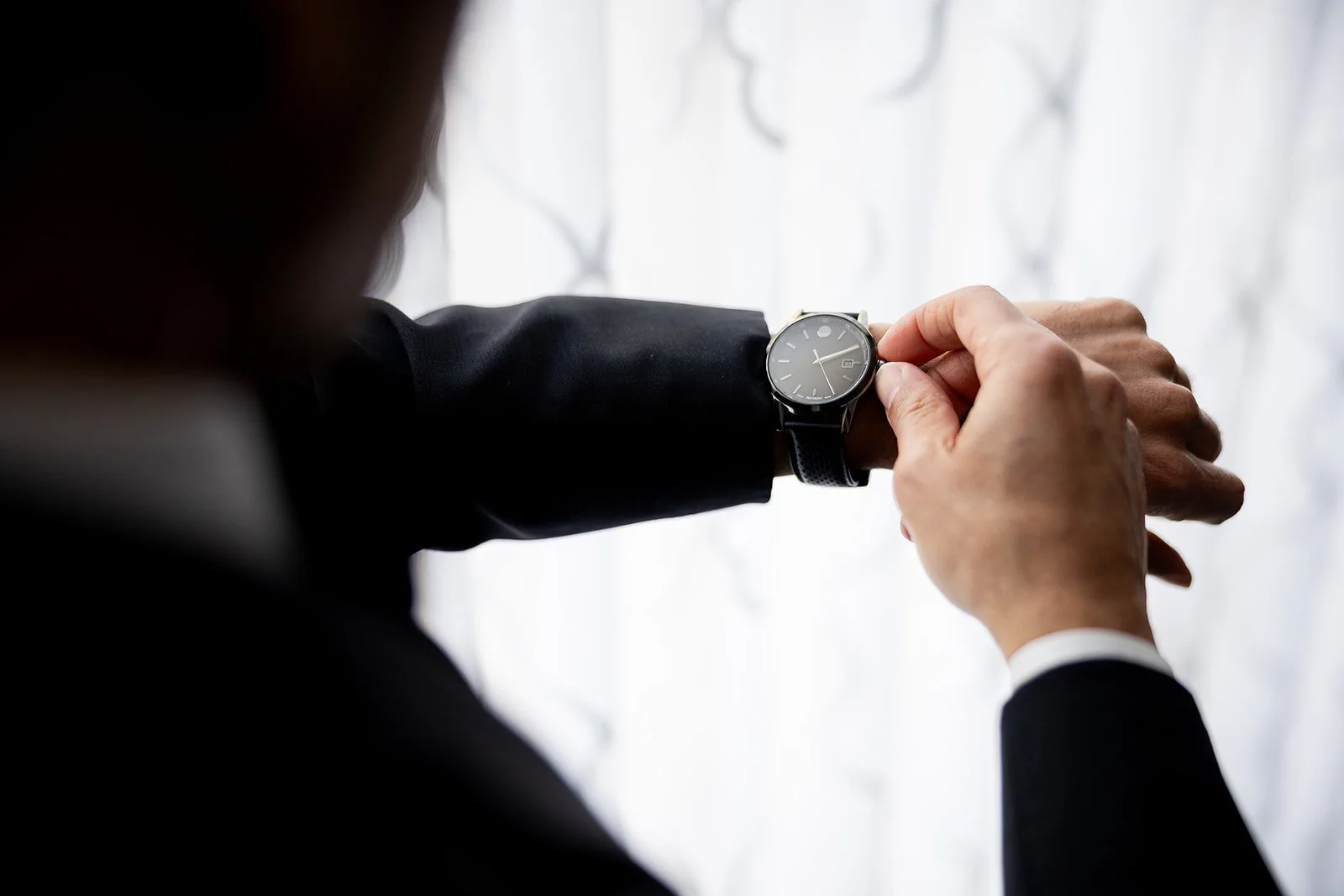 Person in a black suit looking at a wristwatch on their left wrist against a bright background.
