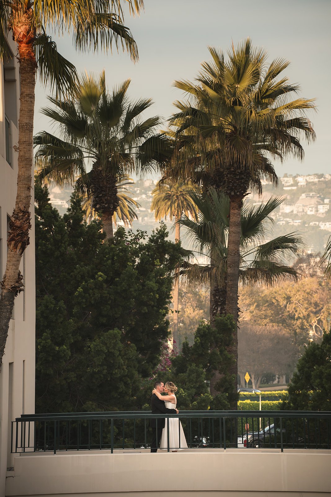 A couple in wedding attire sharing a kiss on a balcony surrounded by tall palm trees