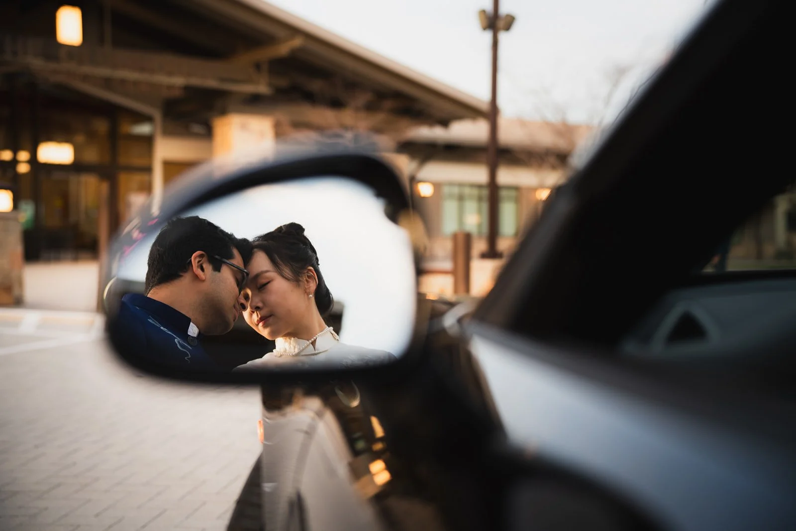 A couple's reflection in a car's side mirror as they lean close together with foreheads touching, outdoors near a building at sunset.