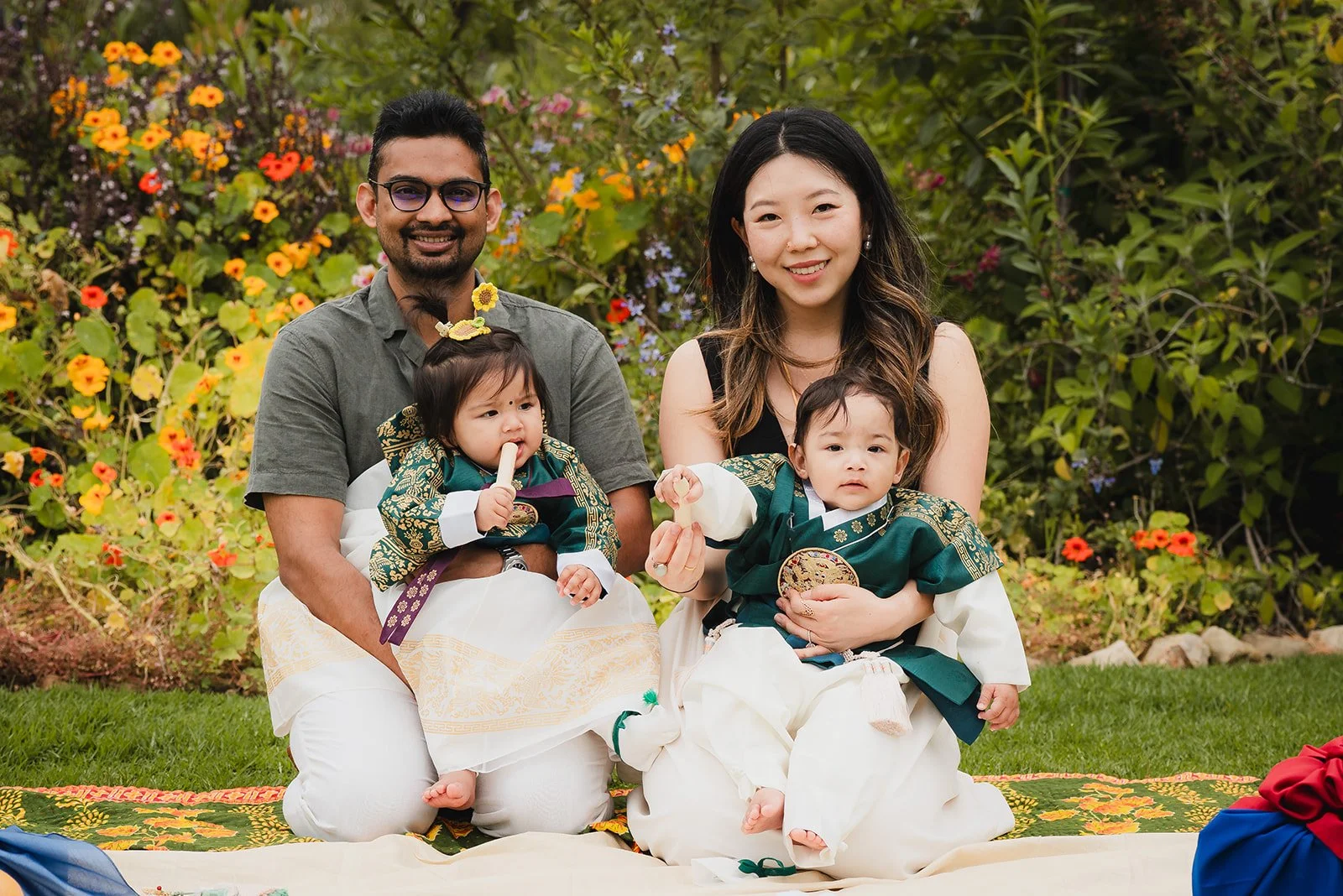 A family of four, including two children, sitting on a colorful mat outdoors in front of flowering bushes. The father has glasses and a beard, the mother has long dark hair, and the children are dressed in traditional clothing, with one eating a snac