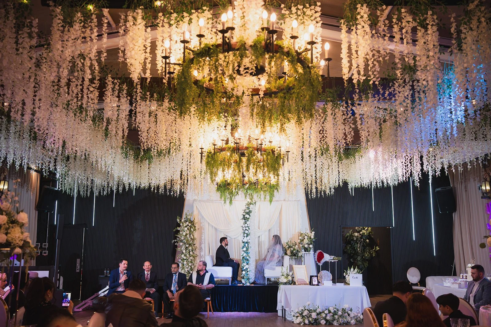 A wedding ceremony with a bride and groom sitting on a platform under hanging white flowers and chandeliers, surrounded by wedding guests in a decorated indoor event space.