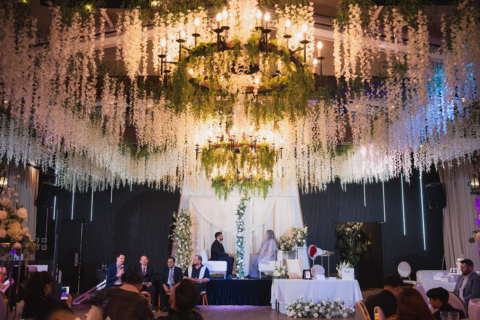 A wedding ceremony with a bride and groom sitting on a platform under hanging white flowers and chandeliers, surrounded by wedding guests in a decorated indoor event space.