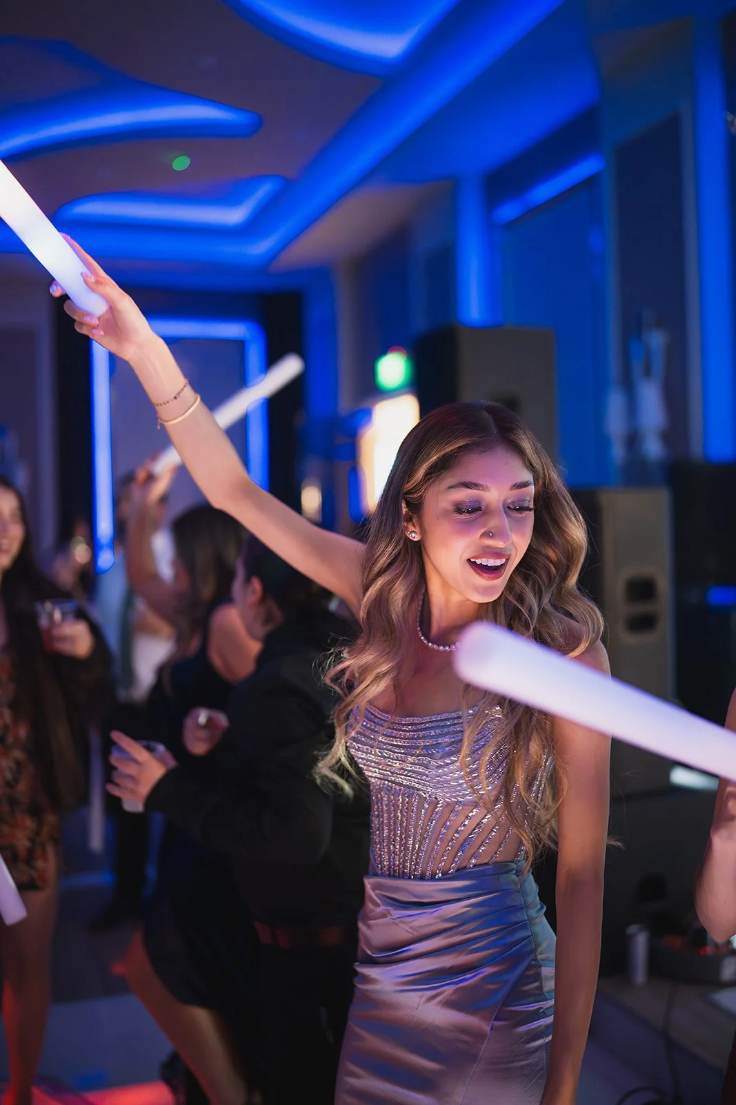 A young woman with long wavy hair dancing at a party, holding a glow stick in each hand, surrounded by other partygoers in a dimly lit room with colorful lighting.
