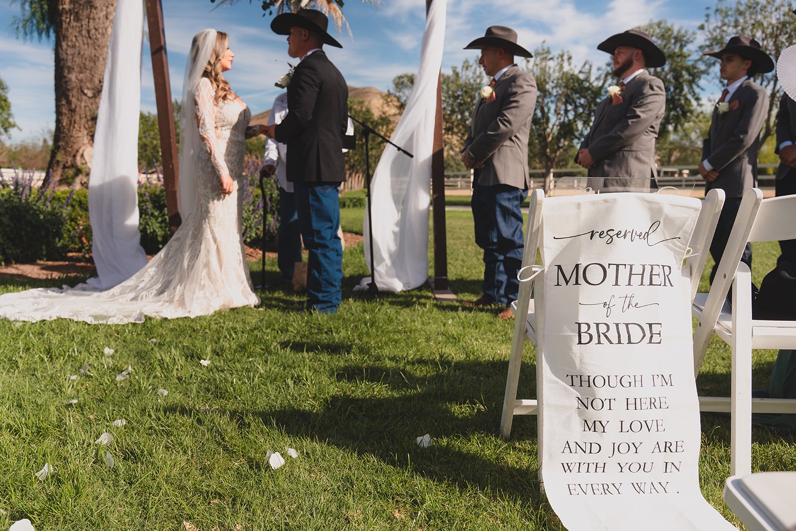 A wedding ceremony outdoors with a bride and groom exchanging vows, surrounded by groomsmen in gray suits and hats. A reserved sign for the mother of the bride is visible in the foreground.