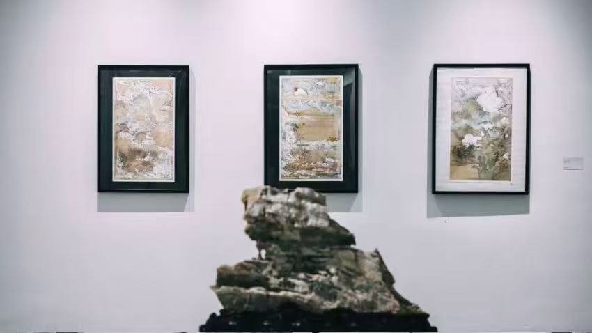 Three abstract landscape paintings in black frames hanging on a white gallery wall, with a textured rock sculpture in the foreground.