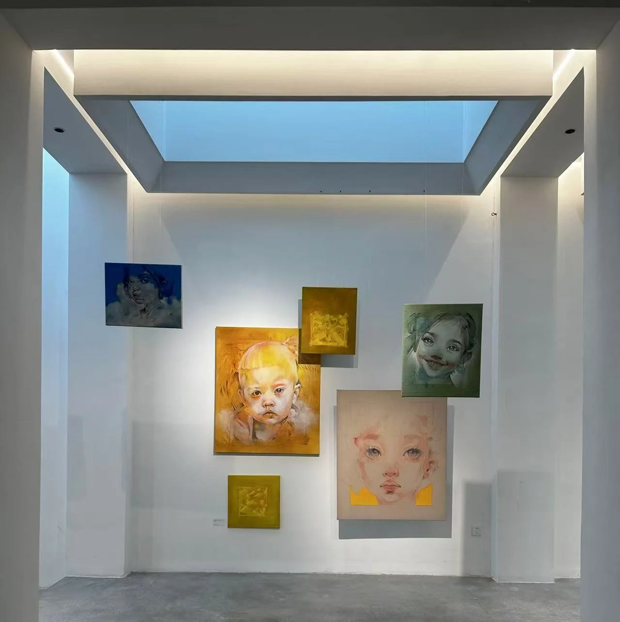 Art gallery display with colorful portrait paintings of children on white walls, illuminated by ceiling lights in a modern space.