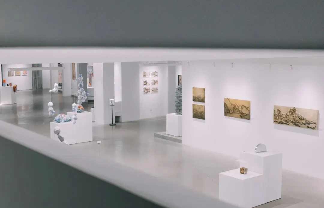 View of an art gallery with various sculptures and paintings displayed on white walls and pedestals, seen through a narrow opening.