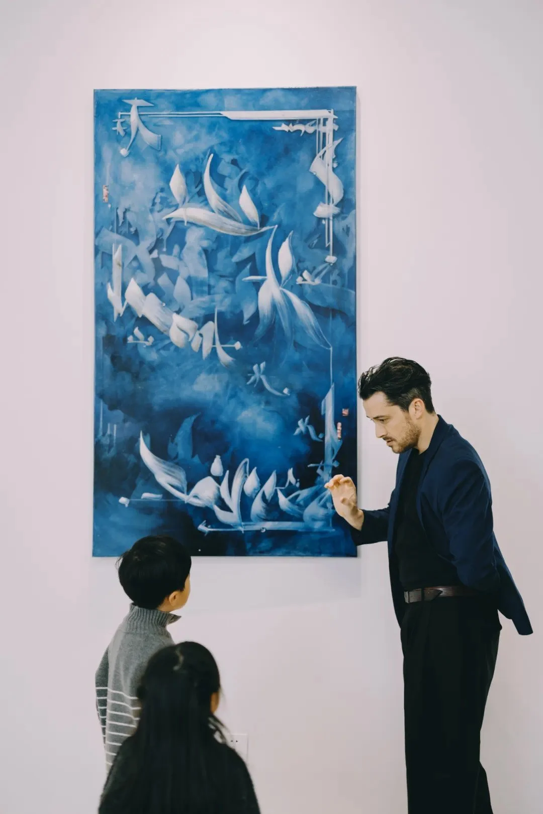 Steve Lecru, 3Dots, Contact page, In an exhibition, explaining artwork to children inside an art gallery. The artwork is an abstract blue painting with flowing shapes.