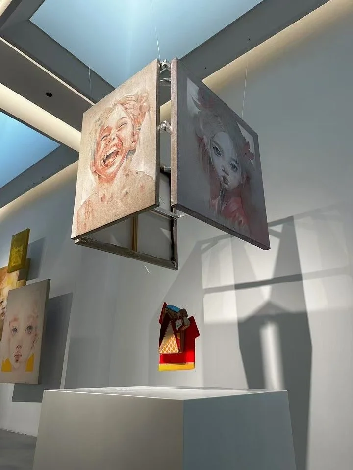 Two paintings of children's faces hanging from a metallic structure in an art gallery, with shadows cast on the wall behind.