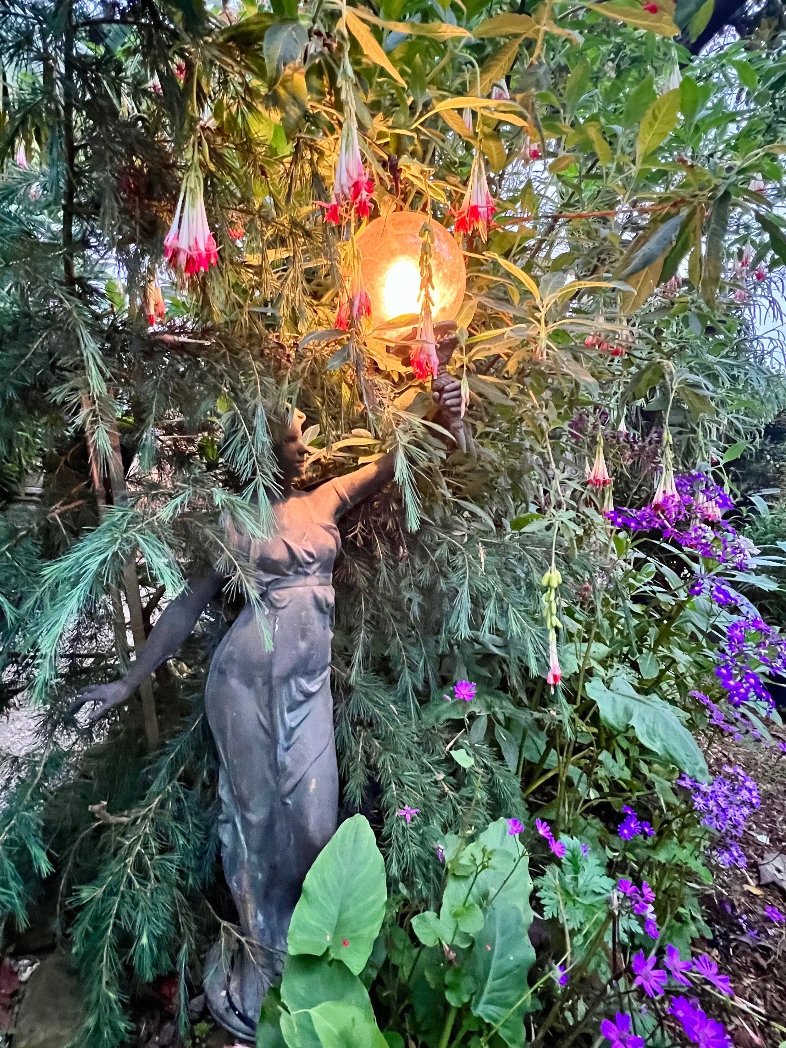 A decorative garden statue of a woman holding a lighted lamp, surrounded by lush greenery and colorful flowers.