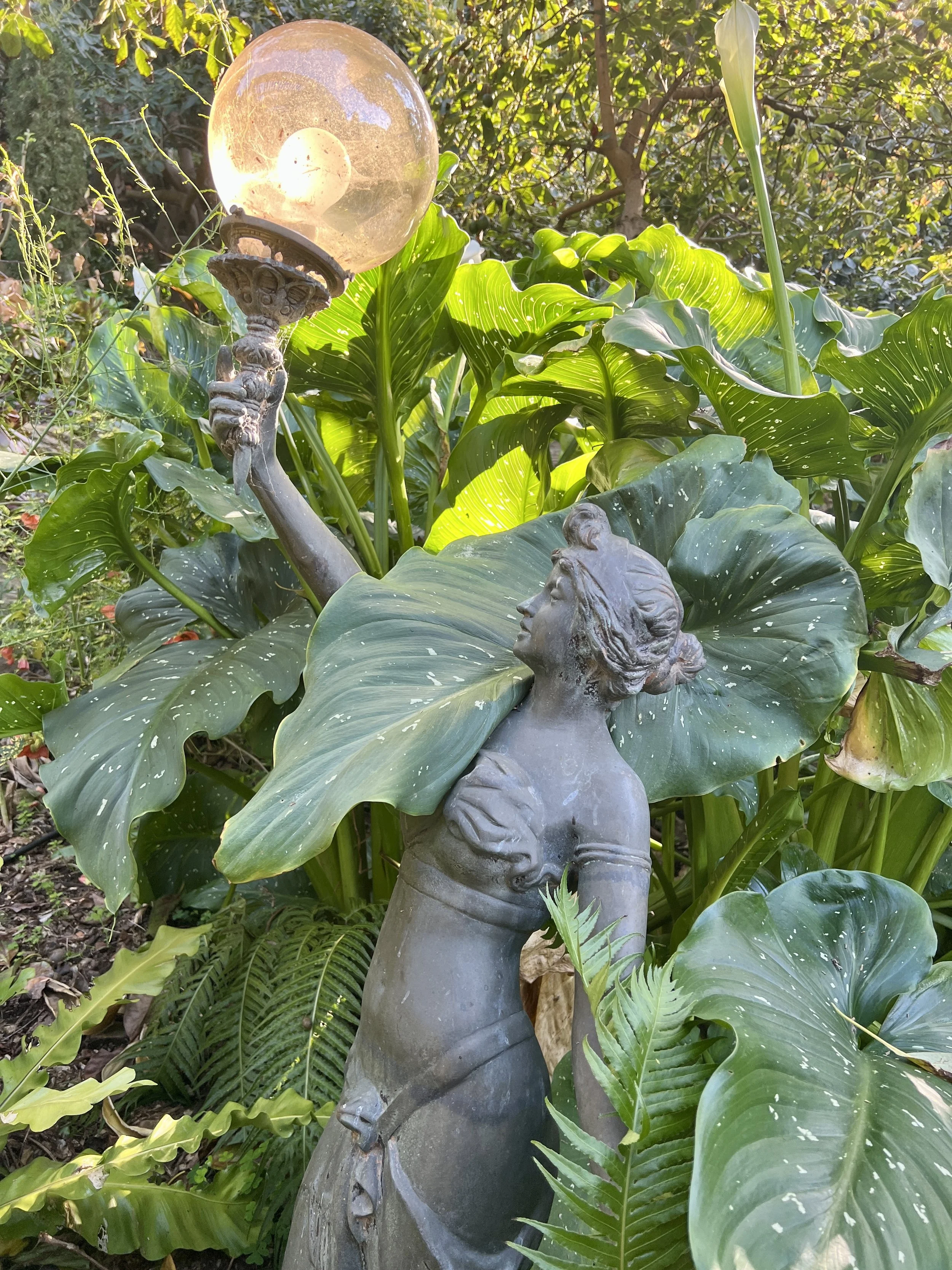 A garden scene featuring a statue of a woman holding a lamp. The statue is surrounded by large green leaves and foliage.