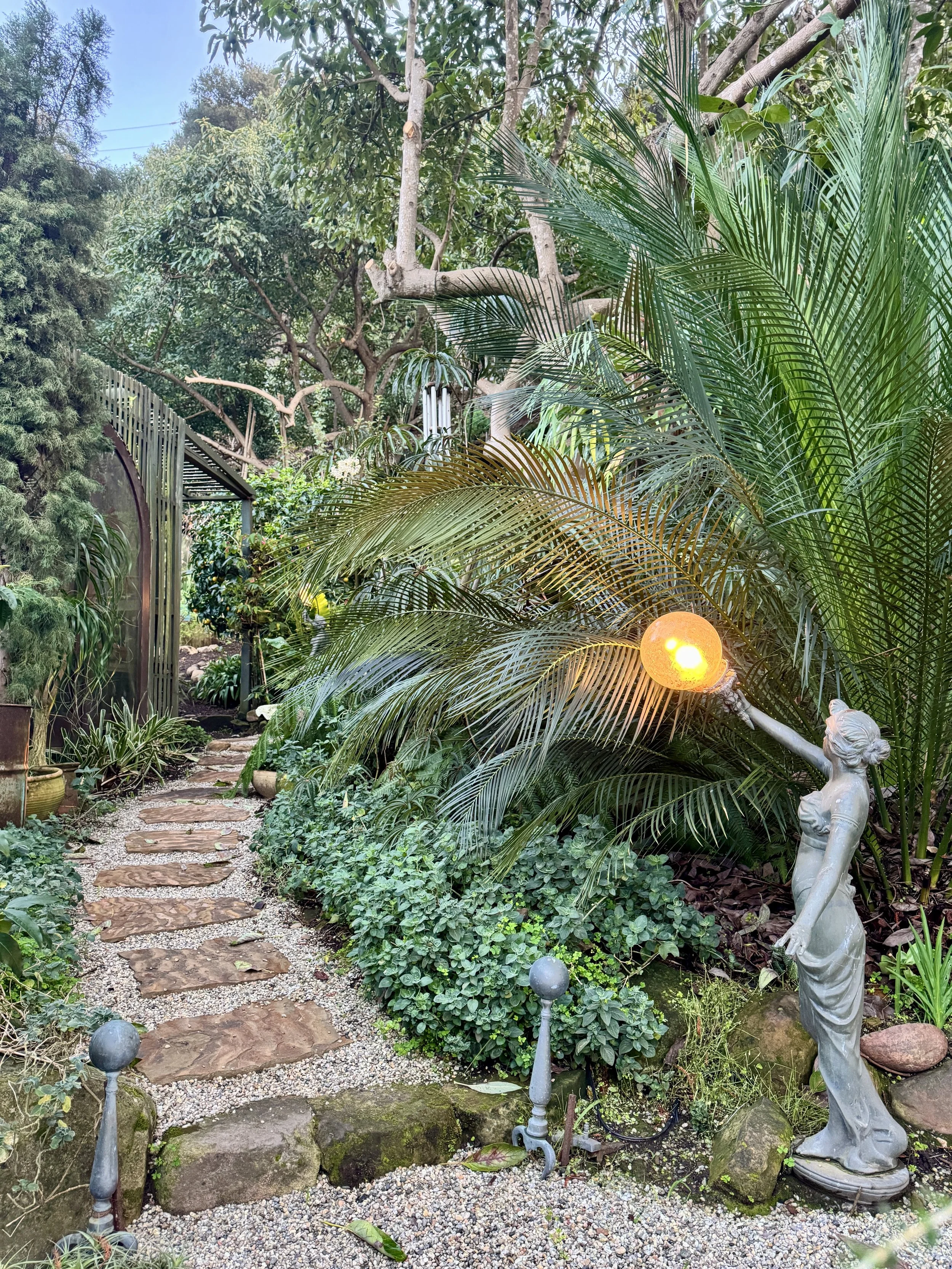 A lush garden pathway with stepping stones, surrounded by green plants and trees, including large palm leaves. A decorative lamp held by a classical-style statue of a woman extends light. Small garden lights line the path.