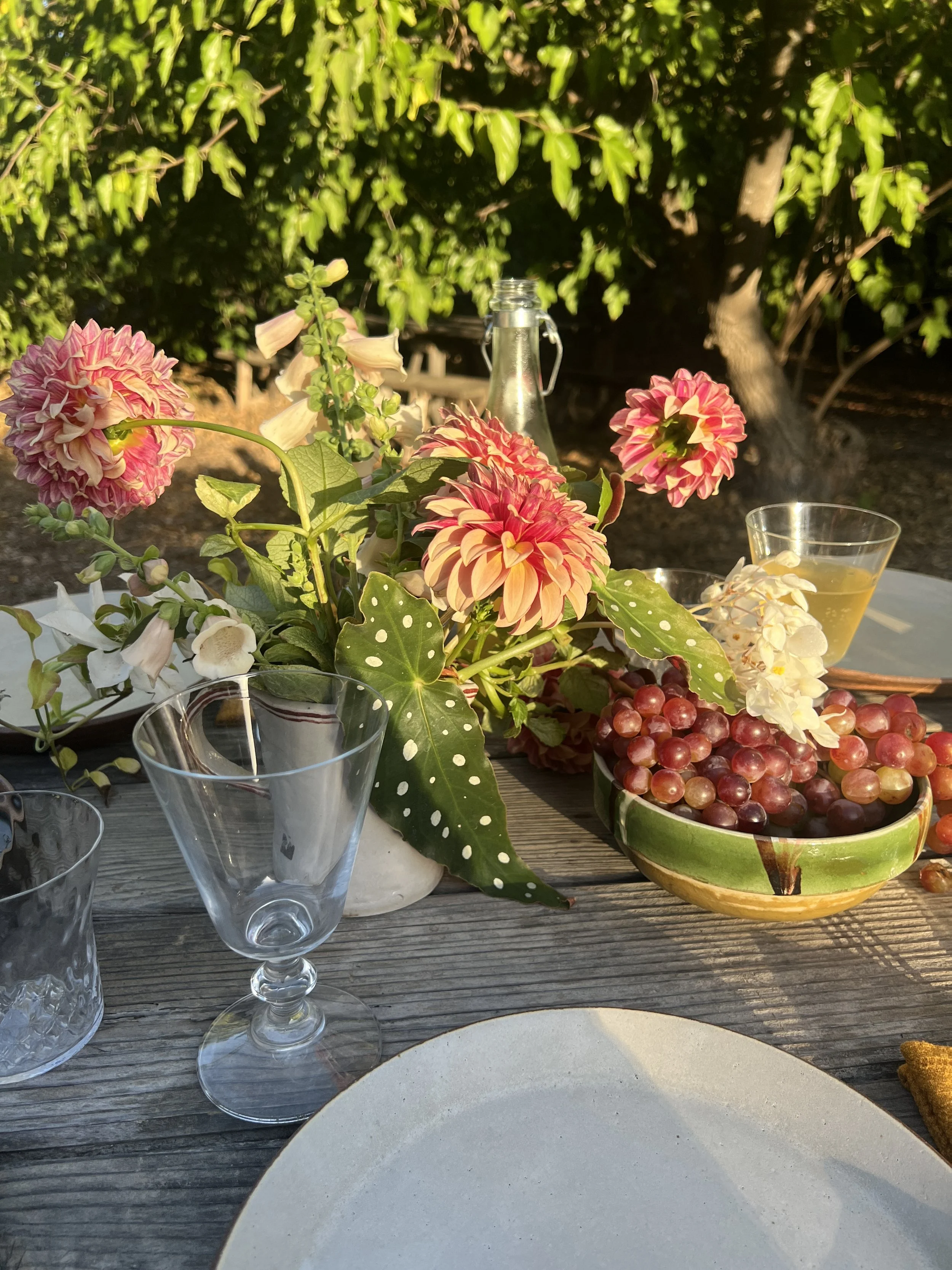 A rustic outdoor table setting with a floral centerpiece, pink and white flowers, a bowl of red grapes, empty wine glasses, and a glass bottle with sunlight filtering through green trees in the background.