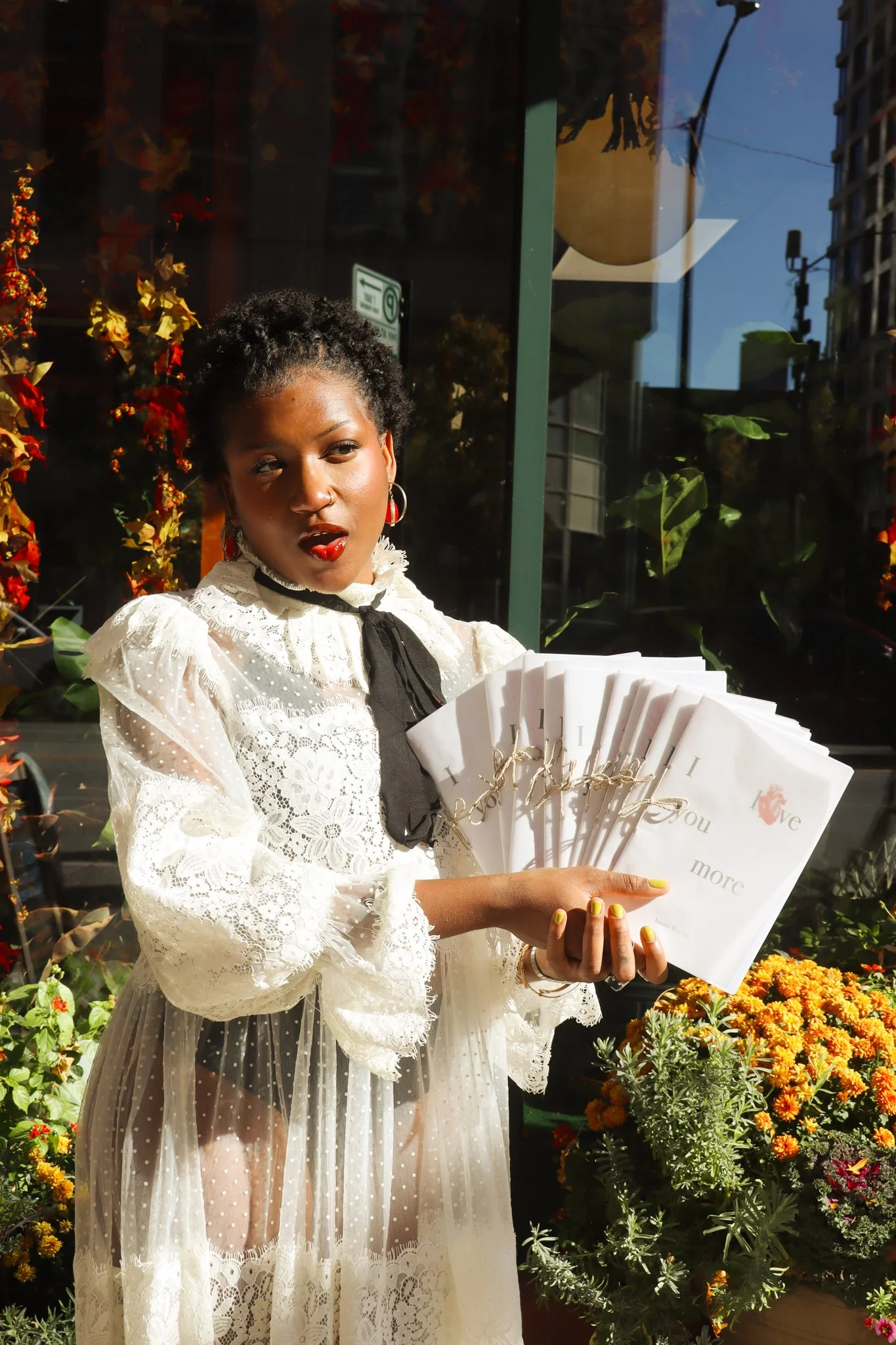 A woman with short curly hair, wearing red lipstick and hoop earrings, stands outside holding several white notebooks with gold writing. She is dressed in a white lace dress with puffy sleeves and a black ribbon tie. There are orange and yellow flowers in front of her and a glass storefront behind her reflecting buildings and street lights.