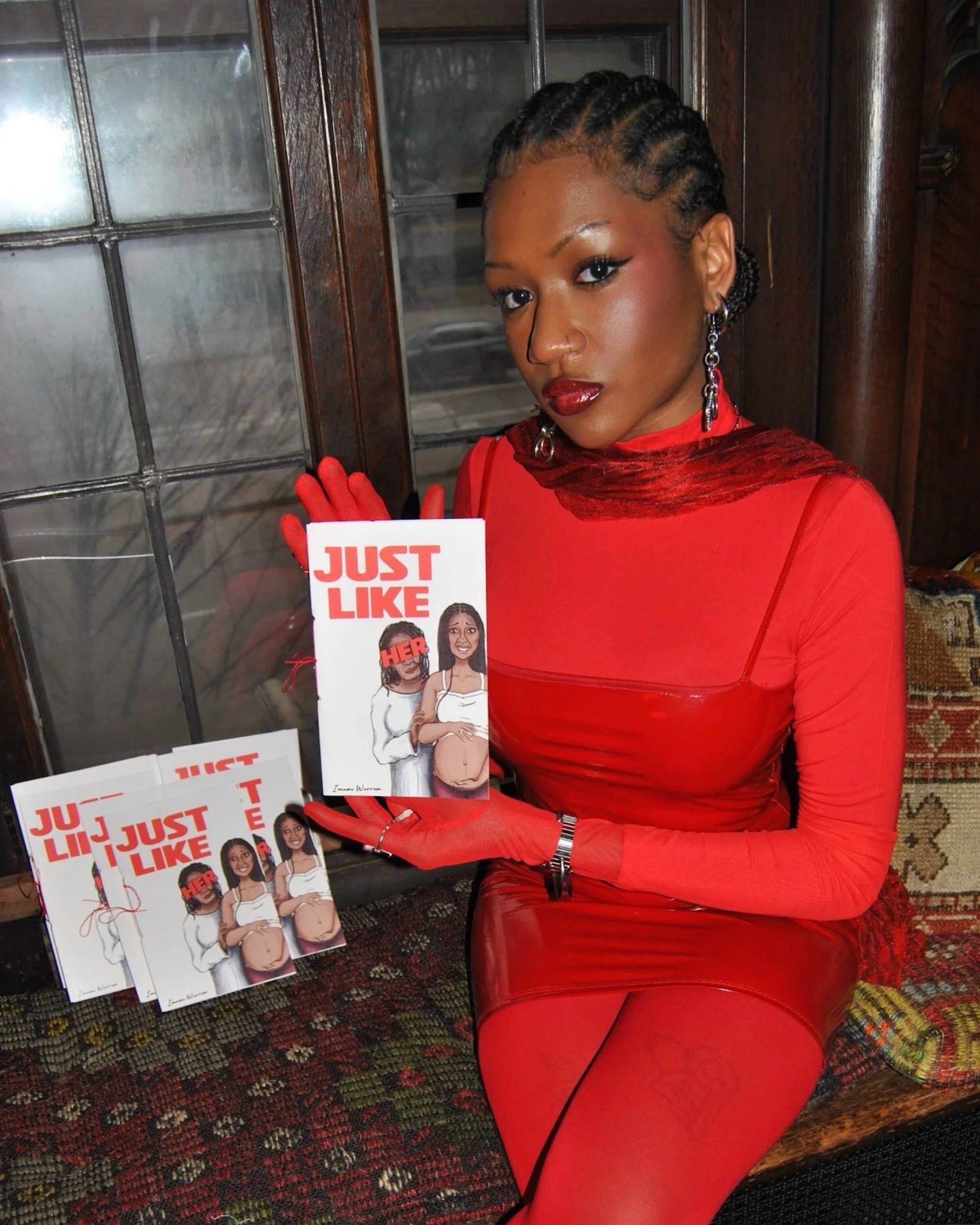 A woman wearing a red dress and matching red gloves sitting on a bench holding a flyer that says 'Just Like Her' with an illustration of a pregnant woman, and several copies of the flyer on the bench.