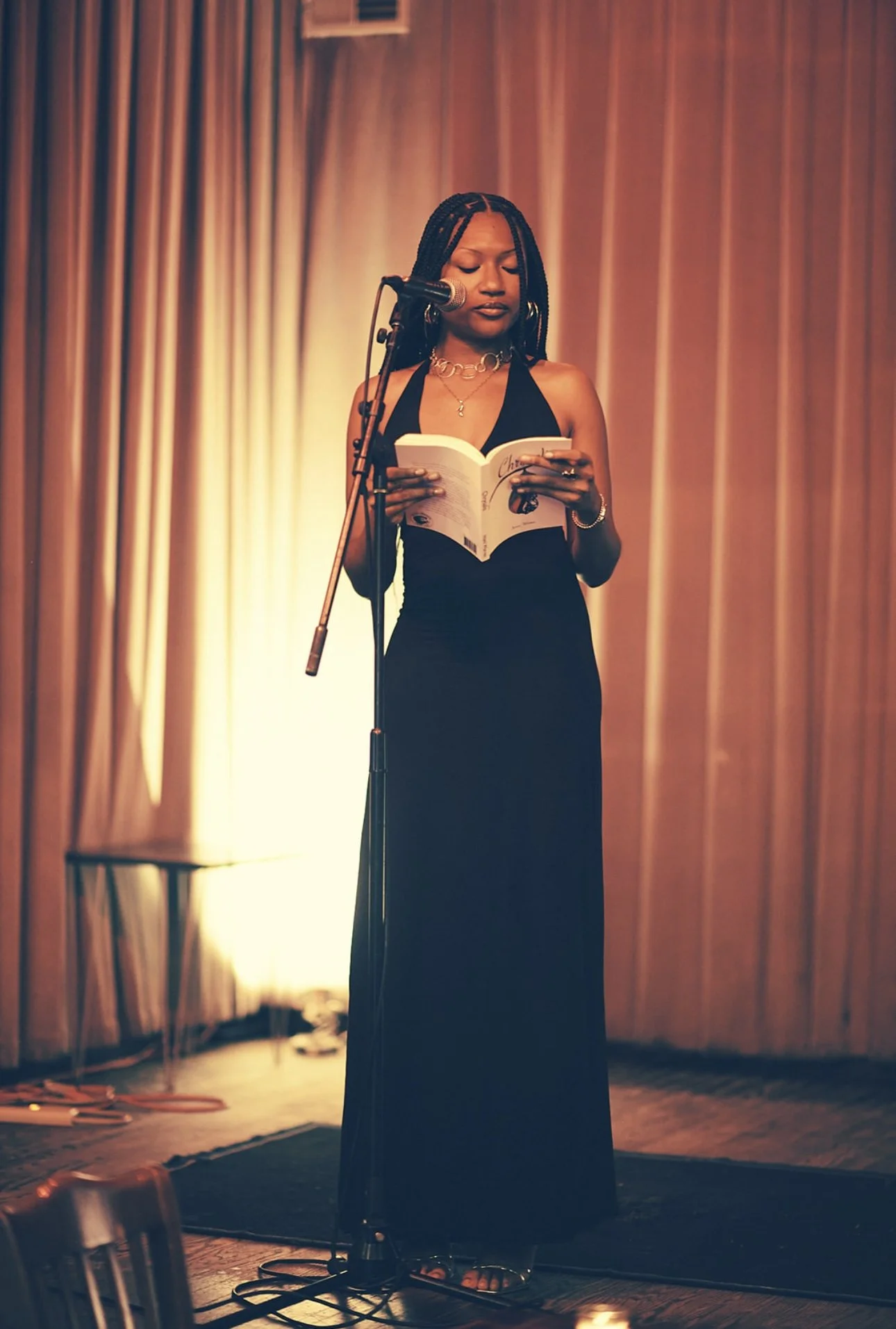 A woman with braided hair, dressed in a long black dress, reading from a book at a microphone on stage with beige curtains in the background.