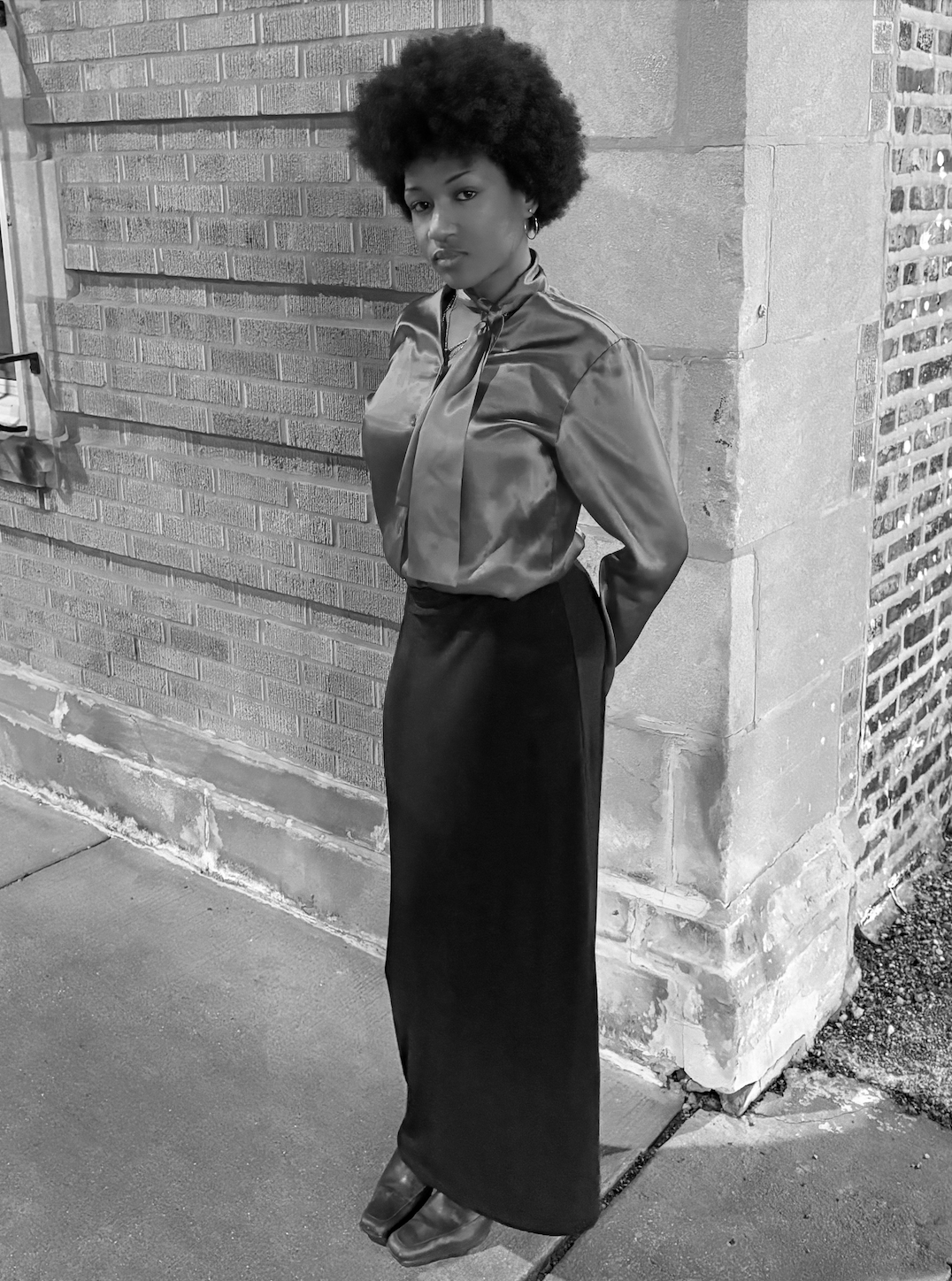 A woman with an Afro hairstyle standing in front of a brick wall in black and white, wearing a shiny satin blouse with a tied neck and a long dark skirt.