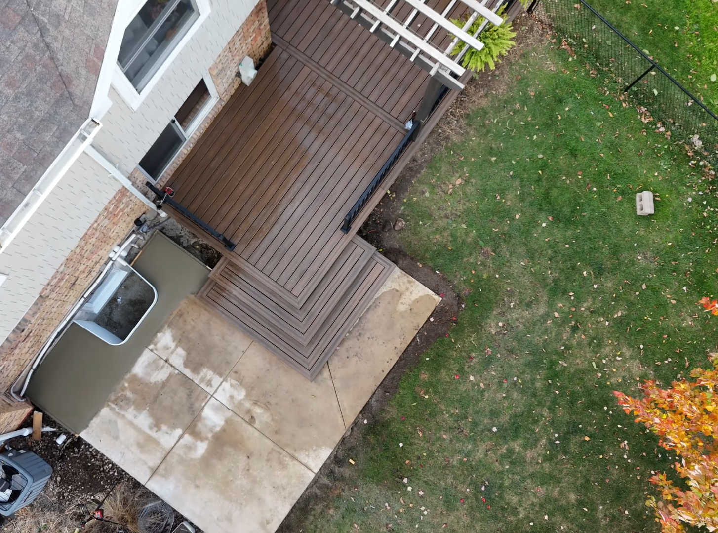 Aerial view of a backyard with a concrete patio, a wooden deck, a green lawn, a small tree with autumn leaves, and a white brick house with windows and an air conditioning unit.