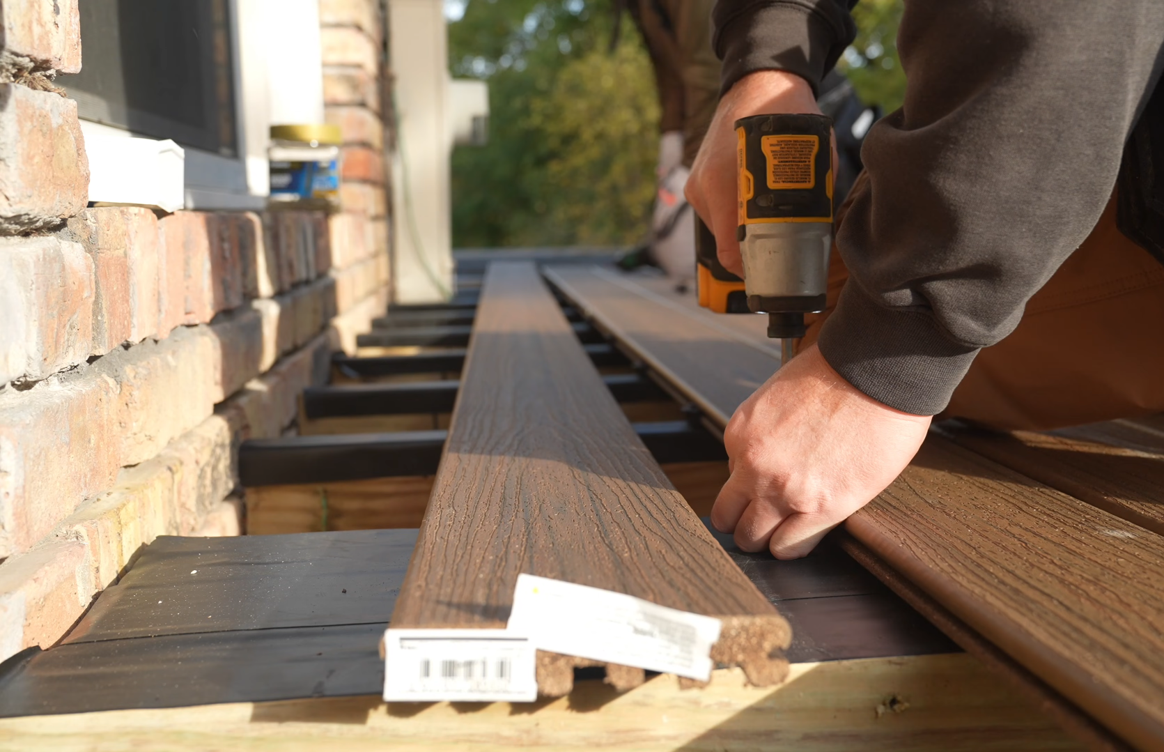 A person using a drill to work on wooden planks on an outdoor construction site, with brick wall and trees in the background.  Carpentyr works.