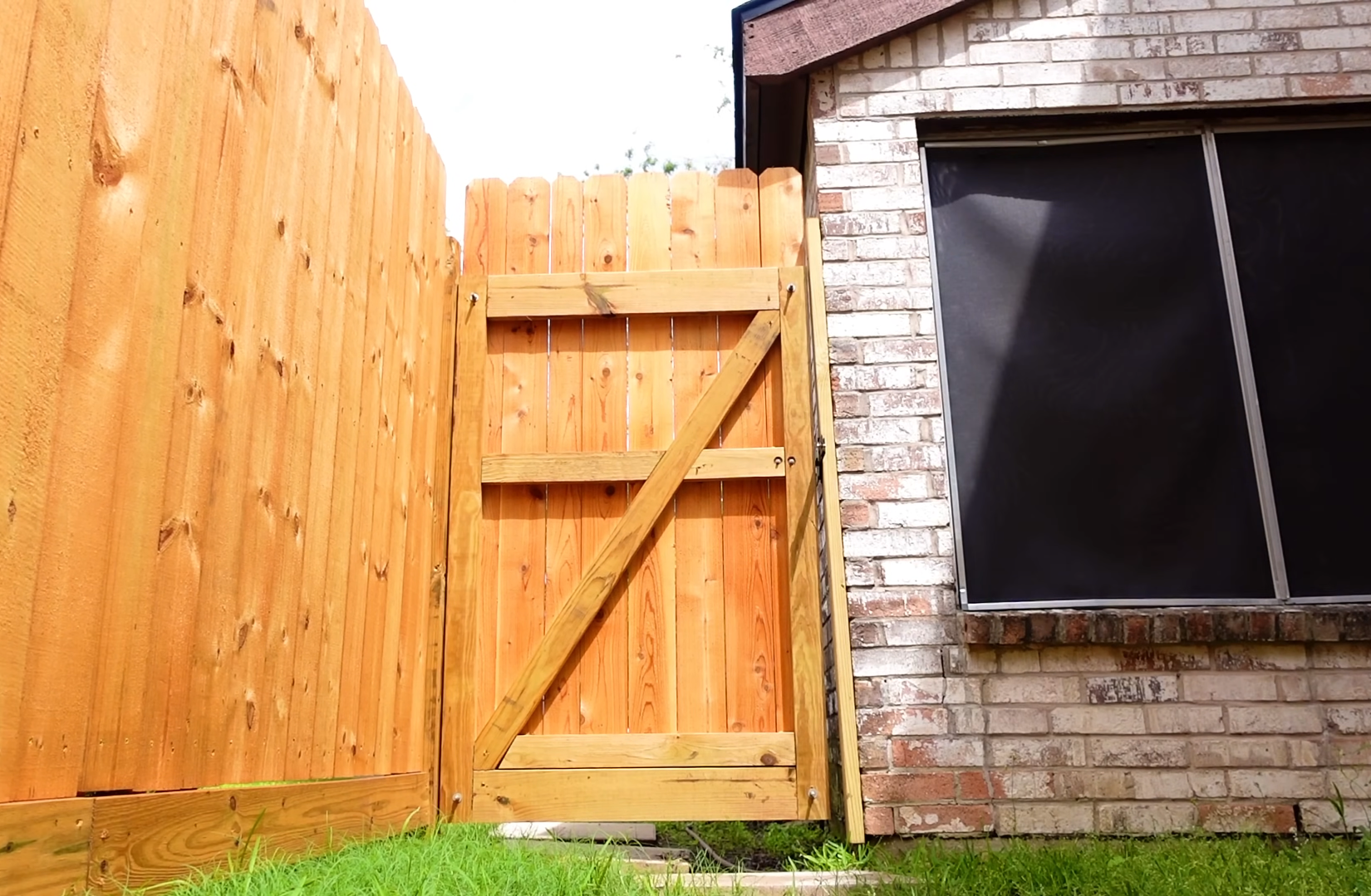 Wooden fence gate attached next to a brick house with a window, green grass at the bottom, and part of the sky visible at the top.
