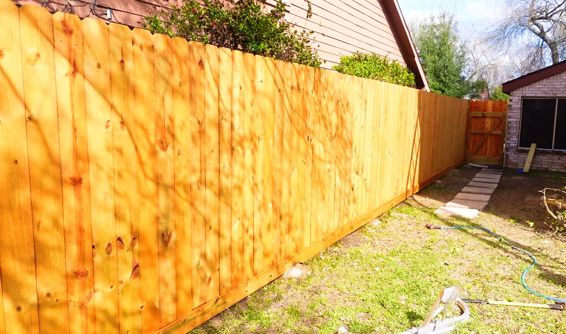 A newly built wooden fence runs along the backyard of a house, with stepping stones leading to a gate. The yard has grass and some construction tools scattered around.