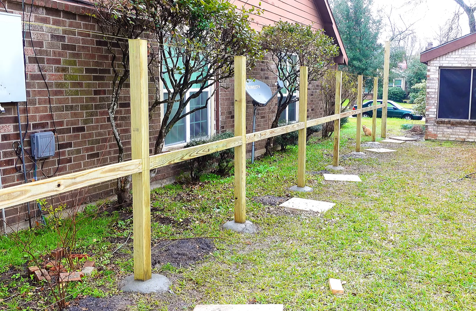 A backyard with a new wooden fence, brick house wall, a satellite dish, and a dog sitting on the grass.