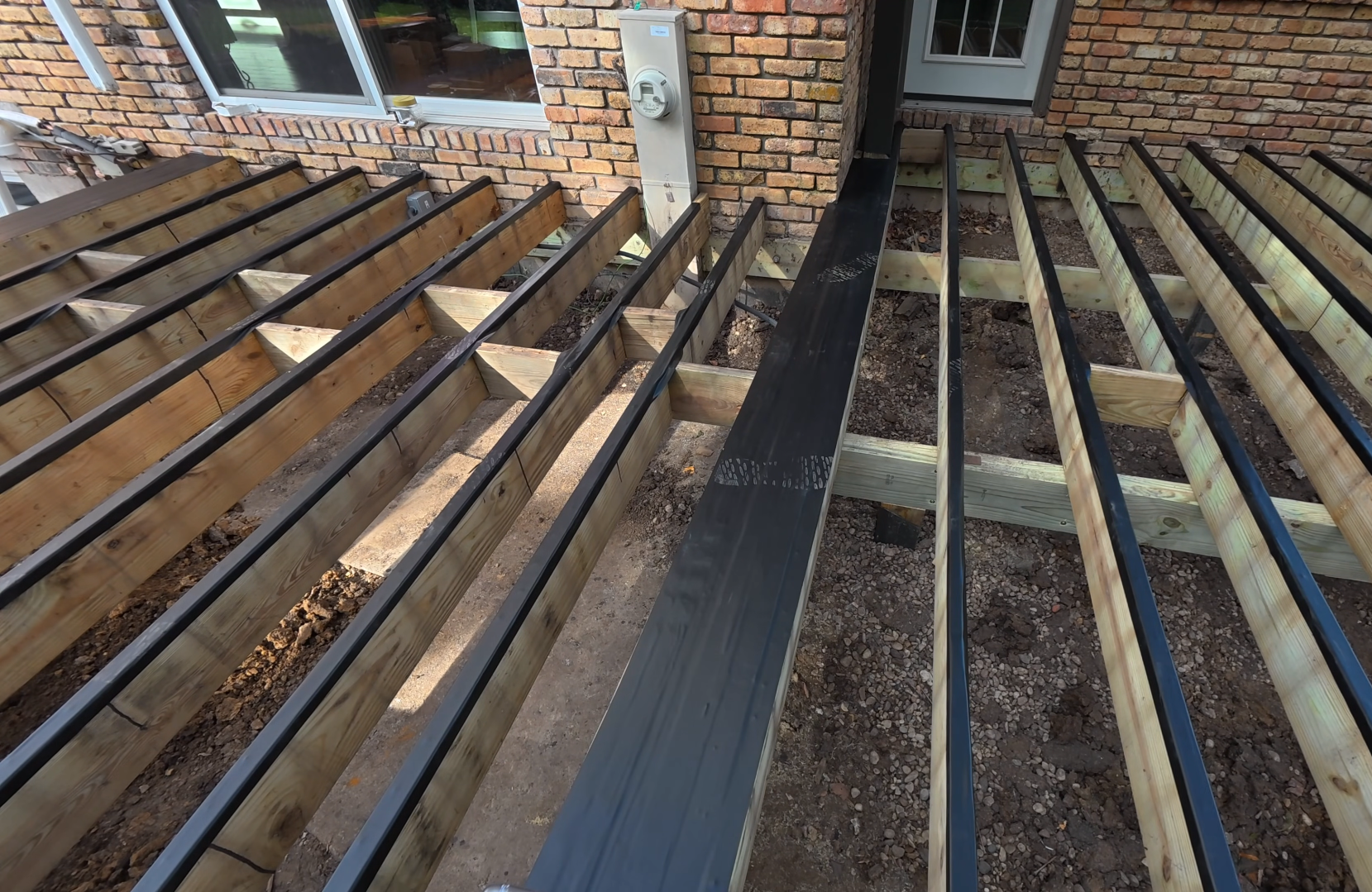 Building deck framing with wooden joists and black metal walkway in progress outside a brick house.