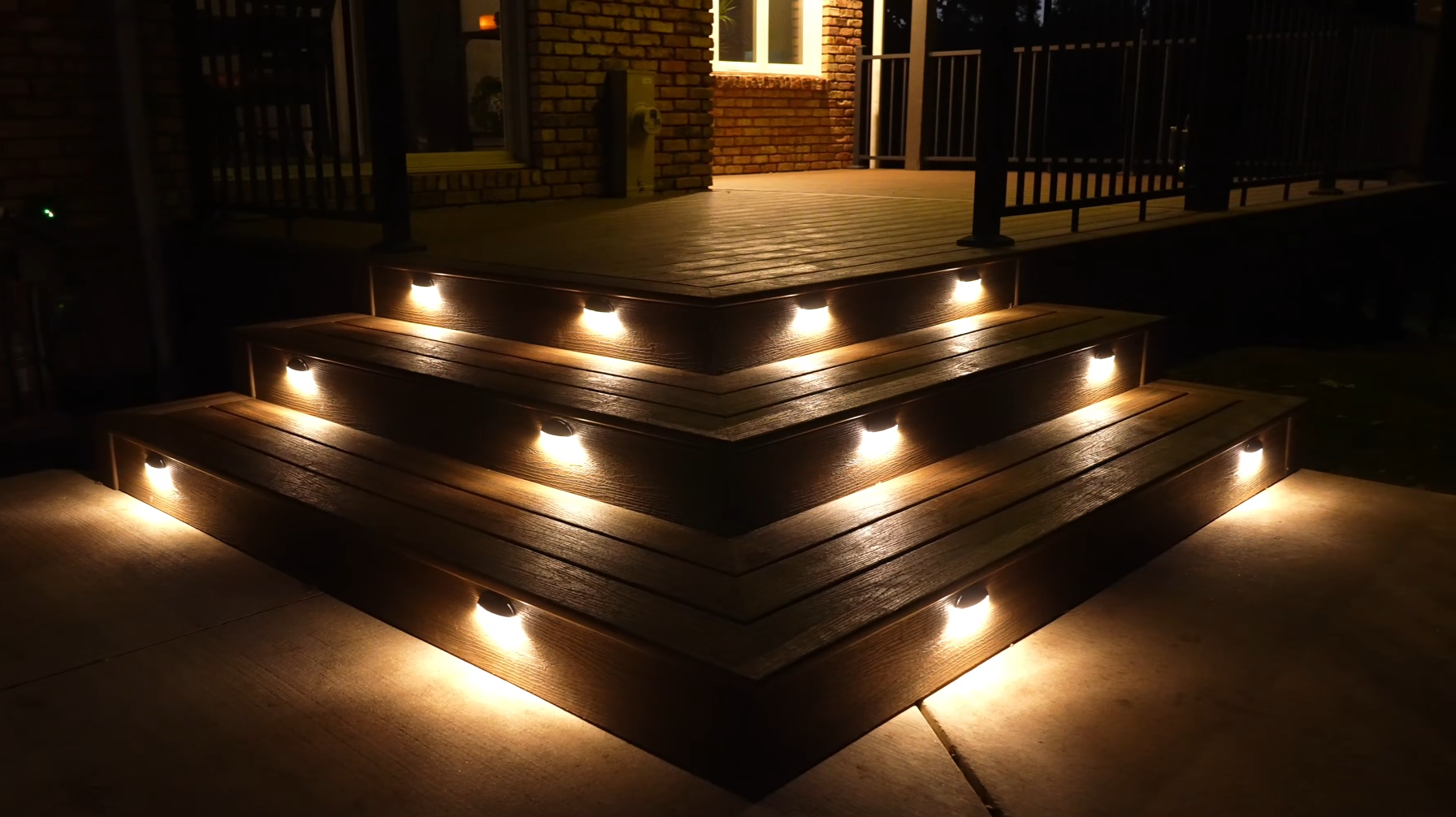 Nighttime photograph of three wooden steps with built-in lights illuminating the staircase leading to a porch of a brick house. Carpentyr works.