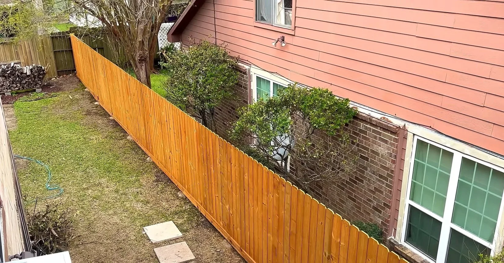 View of a backyard with a wooden fence, grass, trees, and a two-story house with brick and red siding walls.