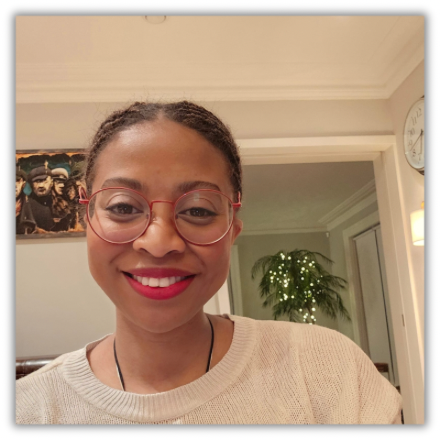 Ifeyinwa Nwaejike, founder and publisher of IFy Atelier, an independent publishing and creative studio in Belfast, Northern Ireland with glasses and red lipstick smiling indoors, with a painting on the wall and a potted plant in the background.