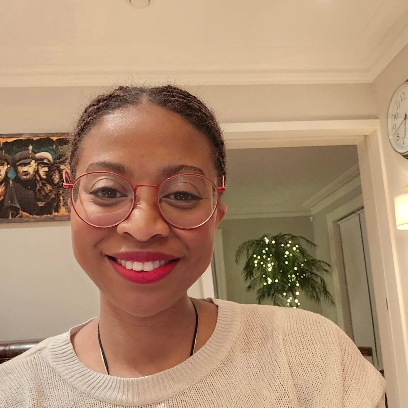 IFy Atelier founder, Ifeyinwa Nwaejike with glasses and red lipstick taking a selfie indoors, with a framed abstract painting, a plant with small white lights, a clock, and decorated walls in the background.