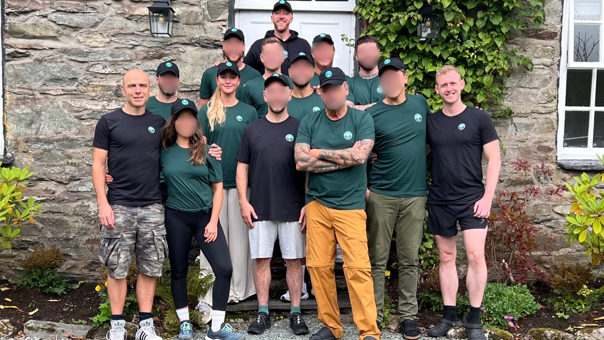 Group of twelve people, some smiling, posing outdoors in front of a stone wall with green plants. They are dressed casually, some in matching green shirts and black caps, others in black or color shorts and T-shirts.