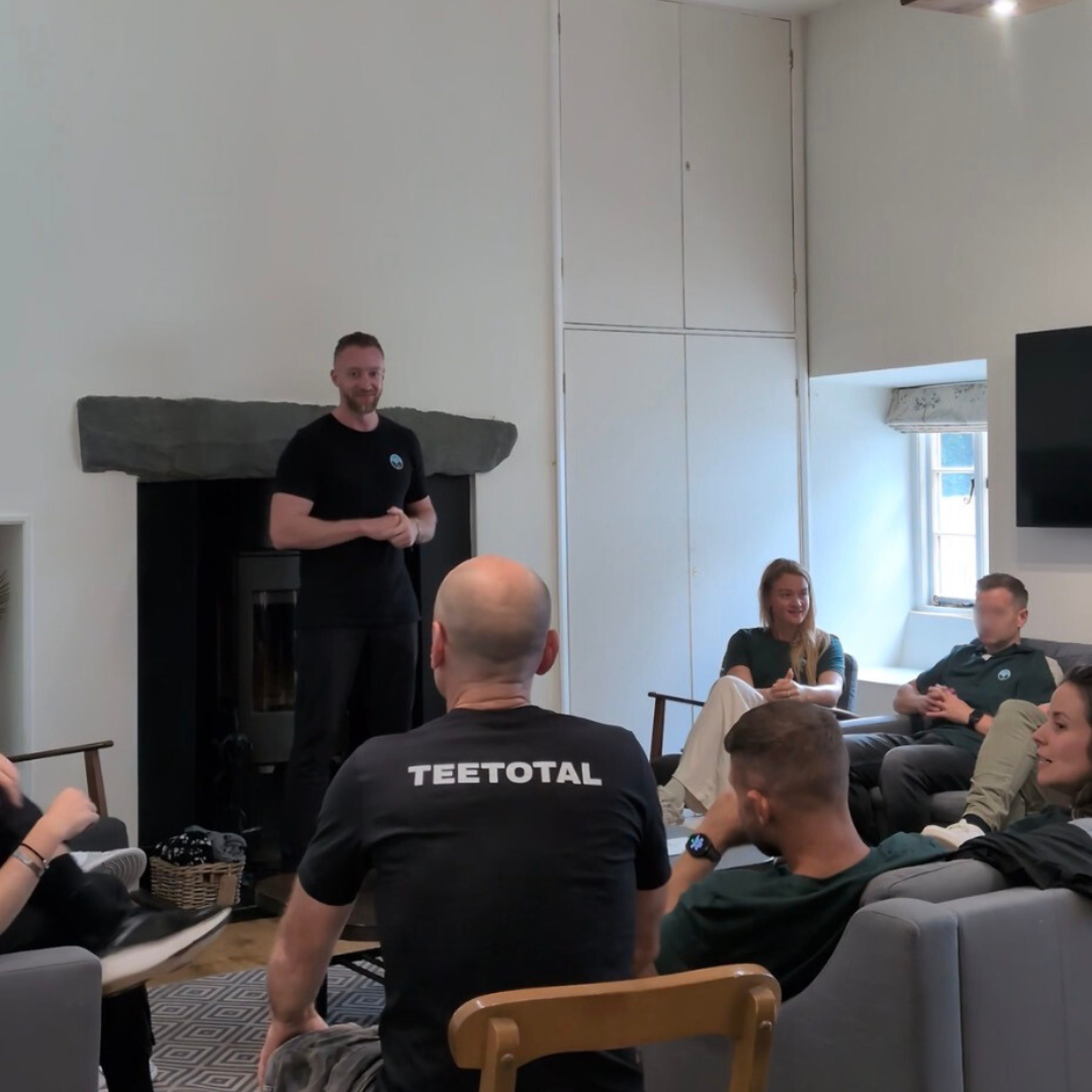 A man giving a presentation to a group of people seated in a cozy room with a fireplace and large windows.