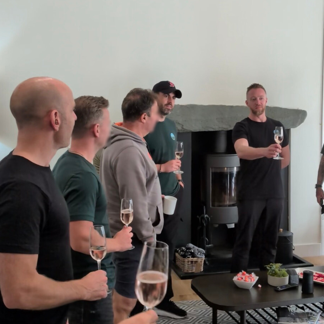 Group of five men standing inside a house, holding glasses of bubbly, with a man on the right giving a toast. There is a fireplace behind them and a table with snacks and drinks in front of them.