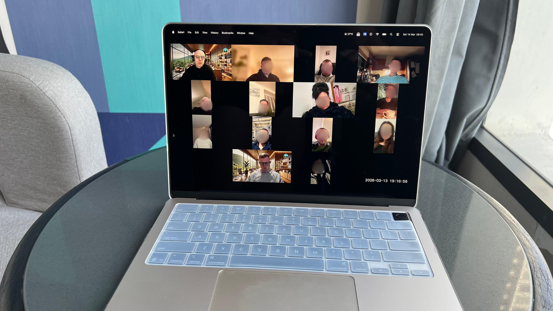 Laptop showing a video conference with multiple participants, sitting on a round table near a window with grey curtains.