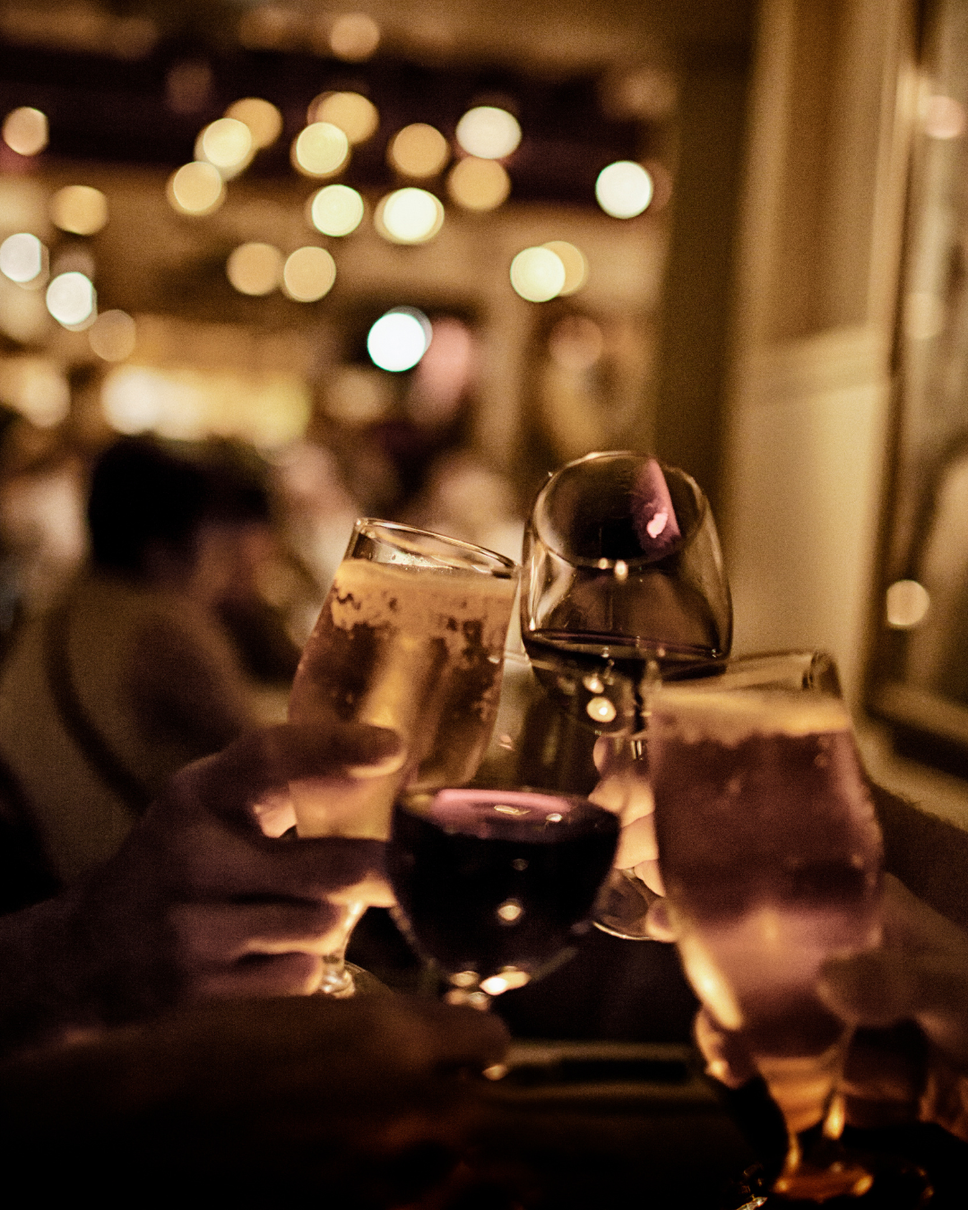 People clinking glasses of red wine and sparkling wine at a restaurant or bar with dim lighting and blurred background.