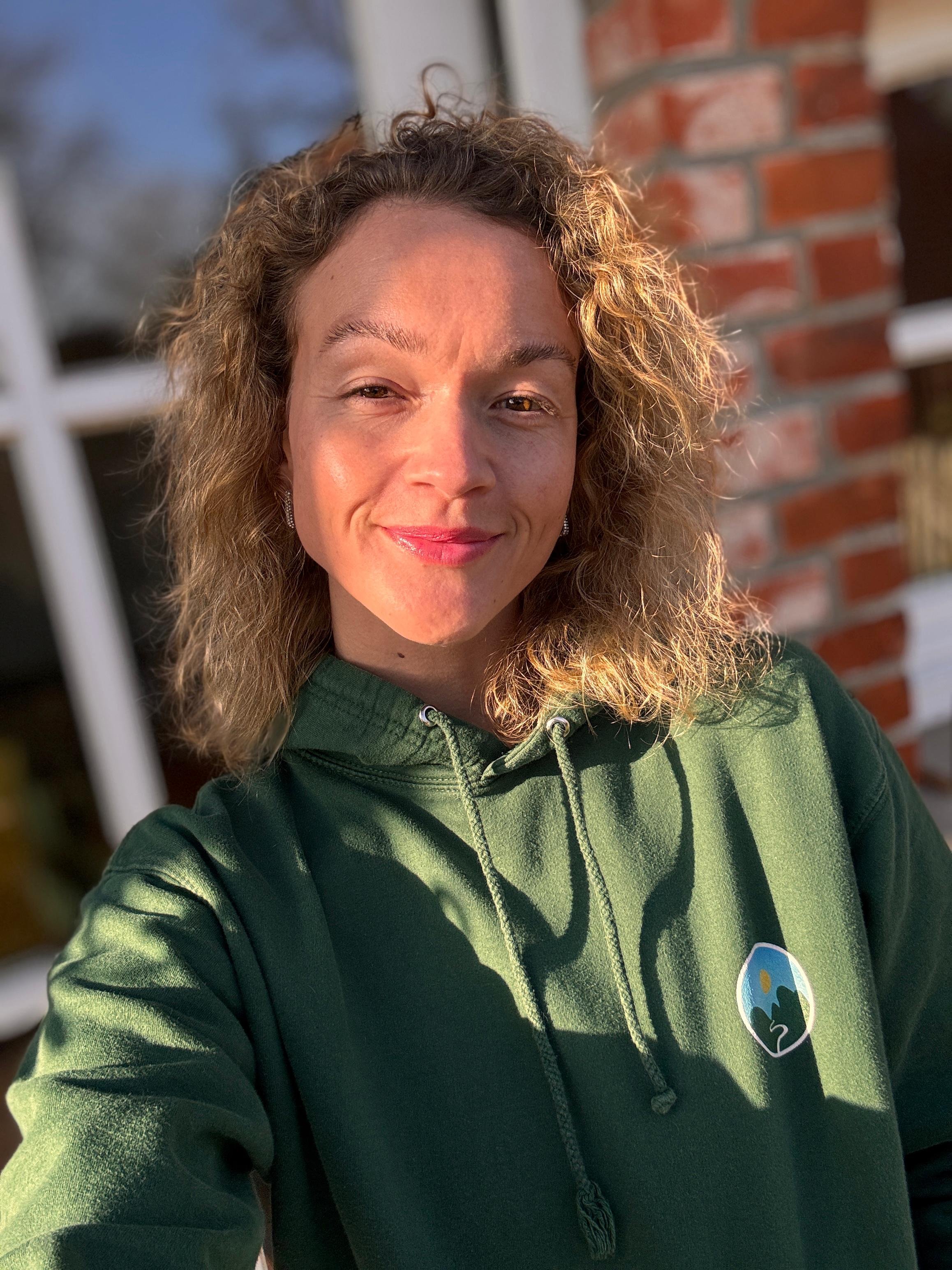 A young woman with curly hair taking a selfie outdoors in sunlight, wearing a green hoodie with a logo, standing in front of a brick wall.