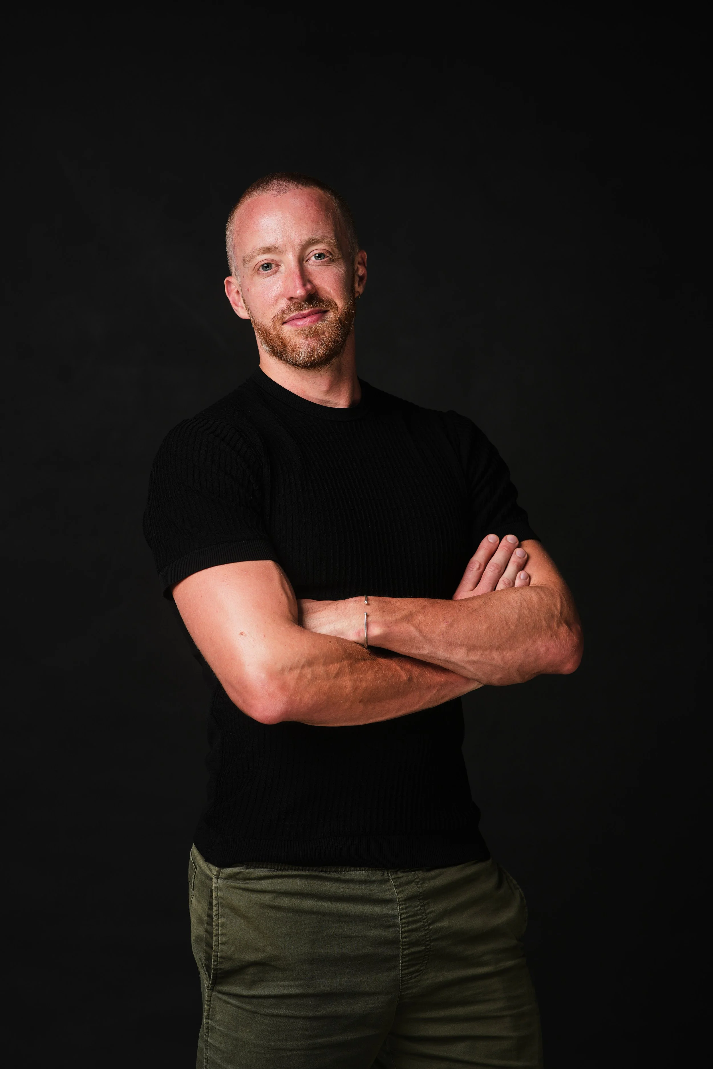 Portrait of a man with short blond hair and a beard, wearing a black short-sleeve shirt and olive green pants, standing with arms crossed against a dark background.