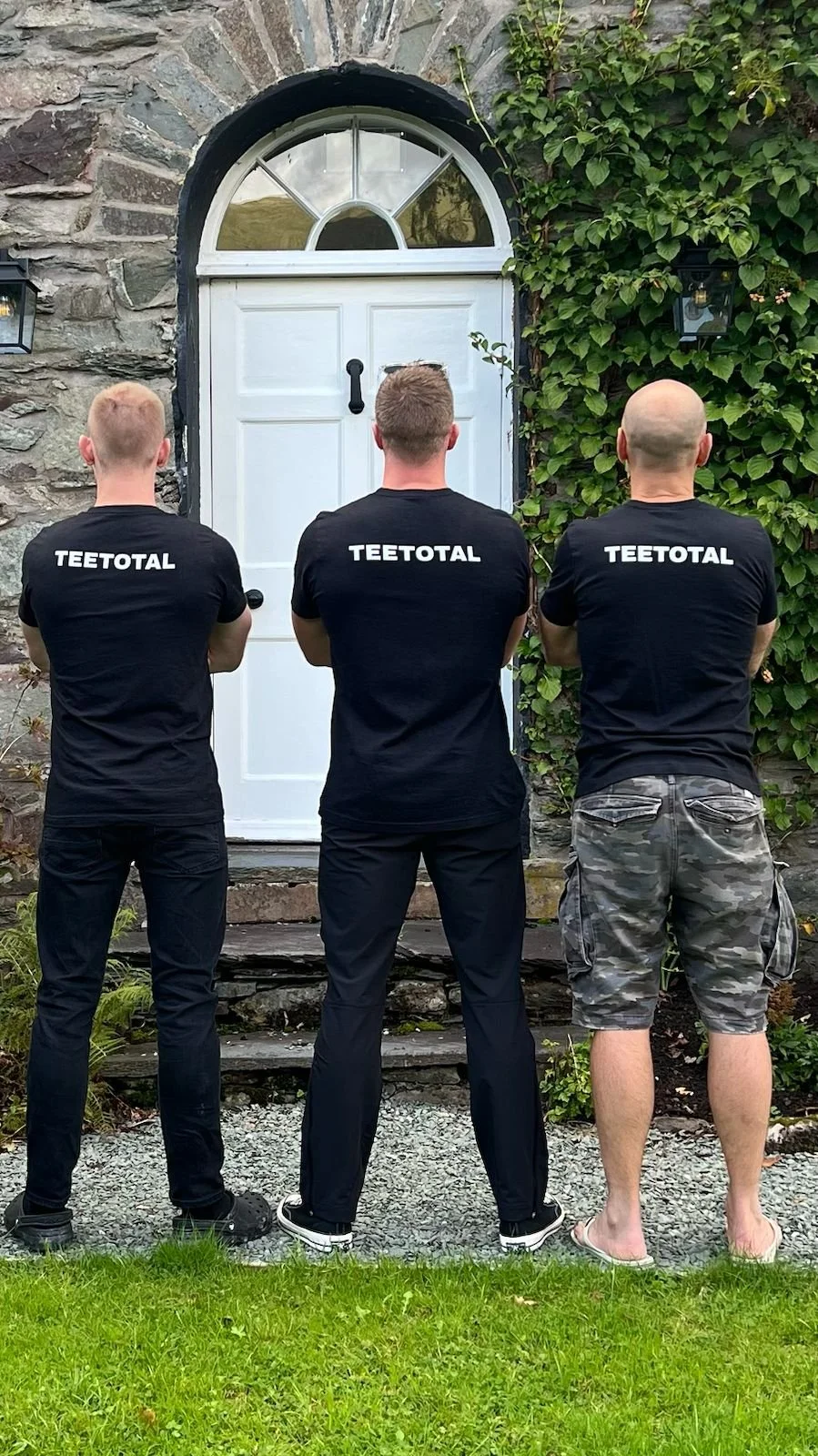 Three men with black t-shirts that say 'TEETOTAL' on the back stand with backs facing the camera, looking at a closed white door with an arched window in a brick wall, surrounded by greenery.