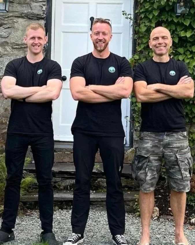 Three men standing outside in front of a white door and stone wall, all wearing black shirts with a small logo and crossing their arms, smiling at the camera.